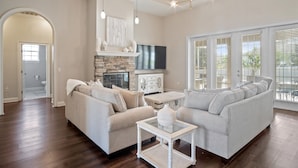 A spacious living room with a stone fireplace, a flat-screen TV, and a large window with blinds.