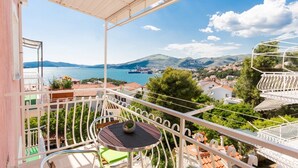 Un balcon avec une table et des chaises, donnant sur une ville côtière et offrant une vue sur la mer et les collines.
