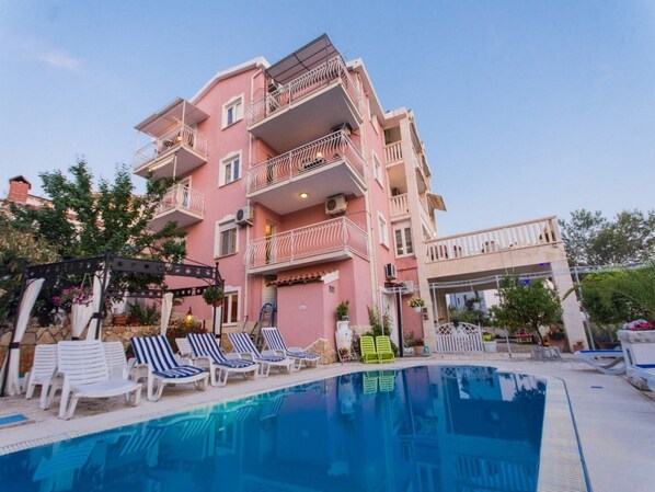 Ein mehrstöckiges, pinkfarbenes Gebäude mit Balkonen, einem Schwimmbad und Liegestühlen.