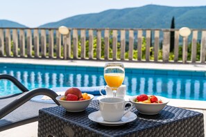Un buffet de petit-déjeuner installé au bord d’une piscine, avec des saladiers de fruits et un verre de jus d’orange.