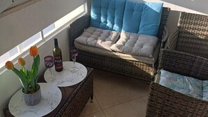 A balcony with wicker furniture, a cushioned seat, a small table with a bottle and glasses, and potted plants. - Apartment Alba - Three Bedroom Apartment with Loggia (Split)