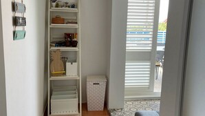 Un pasillo estrecho con un estante blanco contra la pared, una cesta de ropa blanca pequeña y un tapete estampado que conduce a un balcón con contraventanas blancas.