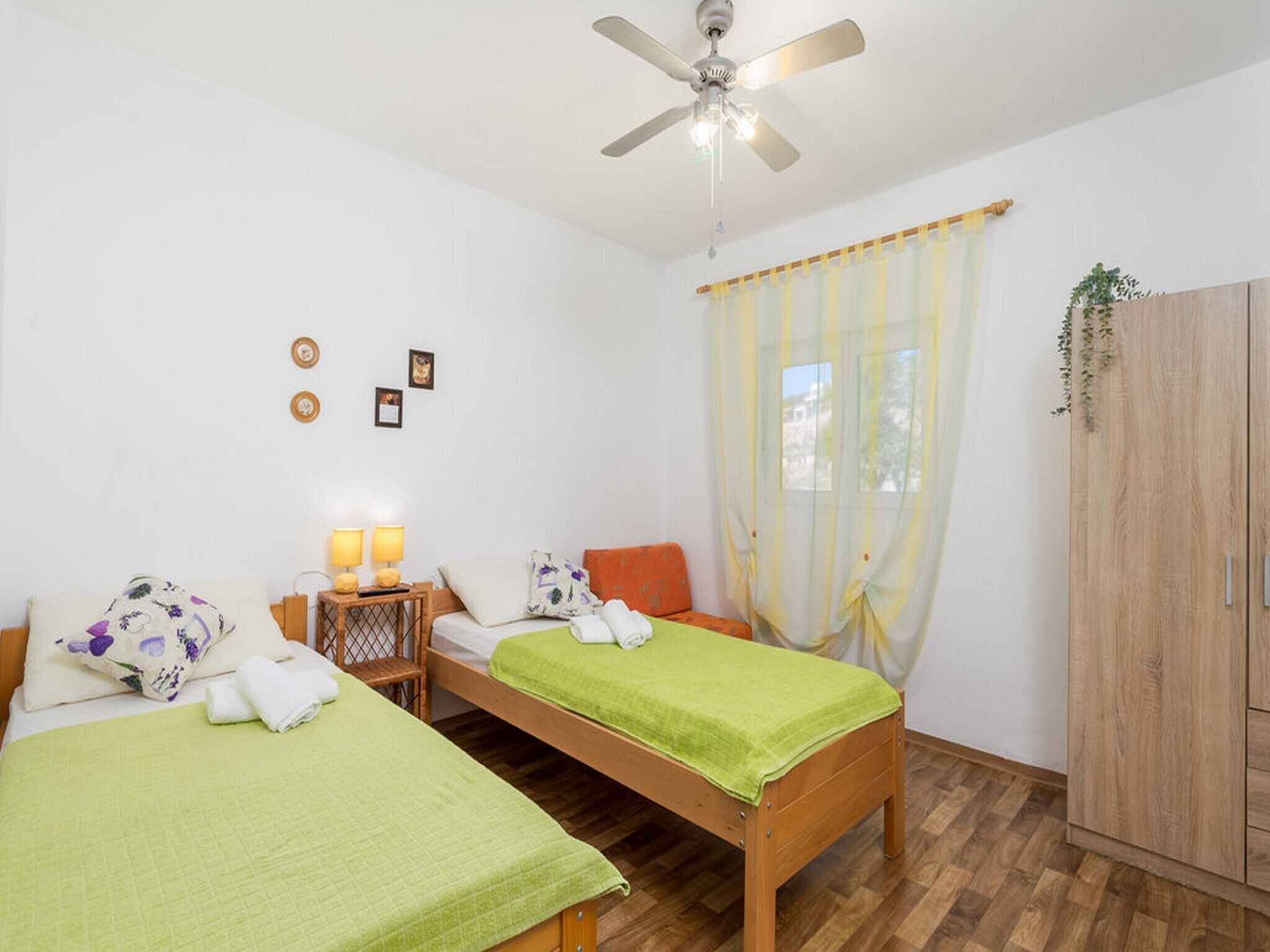 Une chambre avec deux lits simples, une armoire en bois, un ventilateur de plafond et une fenêtre avec des rideaux.