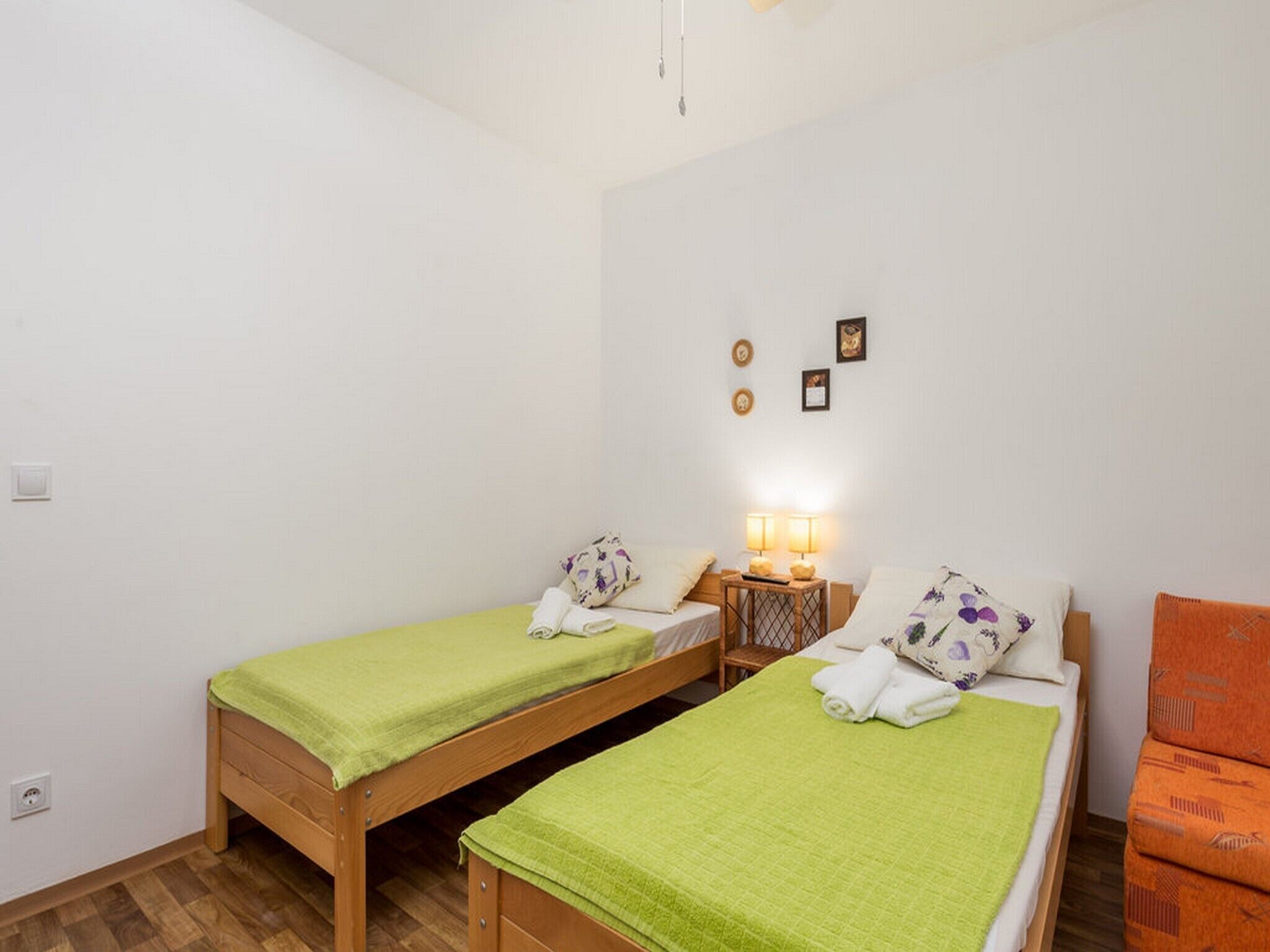 Ein kleines, sauberes Zimmer mit zwei Einzelbetten, einem Holfnachtkästchen mit Lampe und einem kleinen orangefarbenen Sessel.