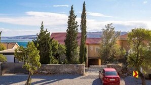 A red car parked in a driveway with a stone wall, trees, and a building with a red roof. - Apartments Sun 4 You - Two Bedroom Apartment with Balcony and Sea View (Karlobag)