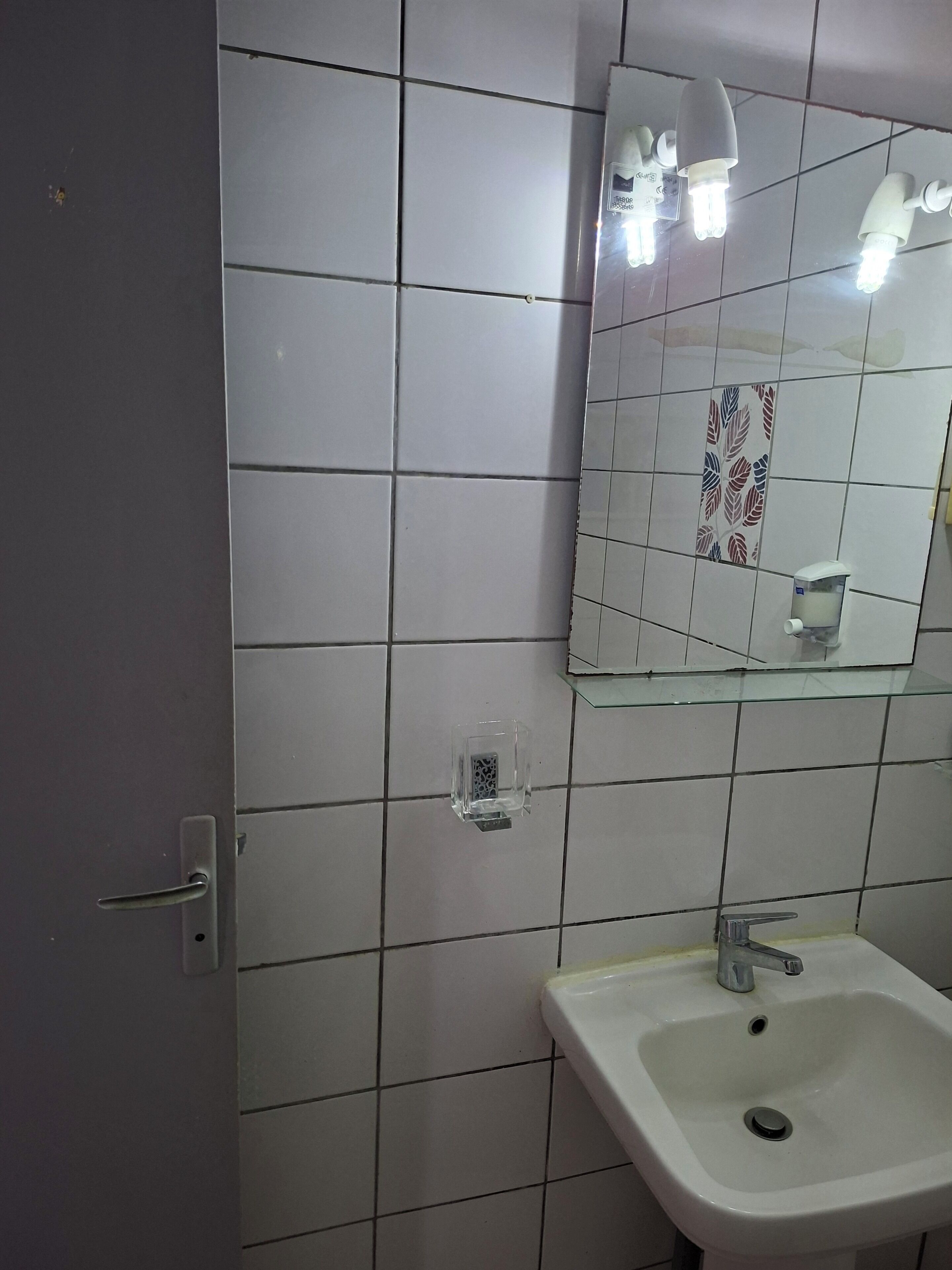 Une salle de bain avec des murs recouverts de carreaux blancs, une étagère en verre, un lavabo blanc avec un robinet et un miroir avec éclairage intégré.