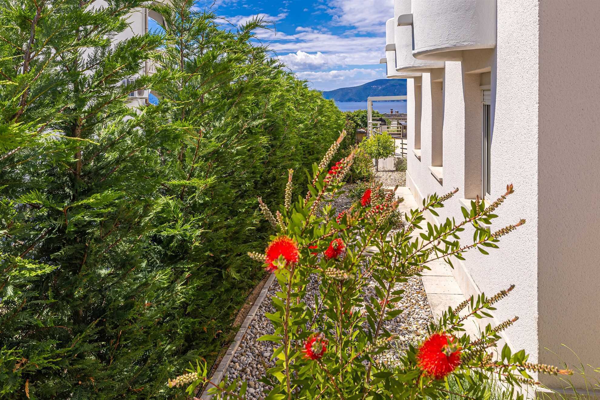 A white building with a balcony overlooking a garden with red flowers and green shrubs.