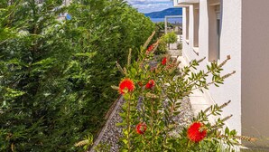 Un edificio bianco con un balcone che si affaccia su un giardino con fiori rossi e siepi verdi.