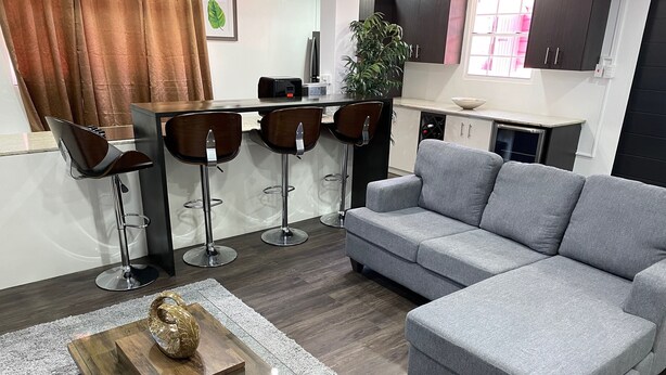 A modern living room with a grey sofa, a wooden coffee table with a decorative item, and a glass-top coffee table. There are three bar stools and a dining table with a plant and a small table lamp.