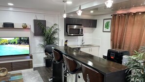 A modern kitchen with a dark countertop, bar stools, a microwave, and a TV displaying a beach scene.