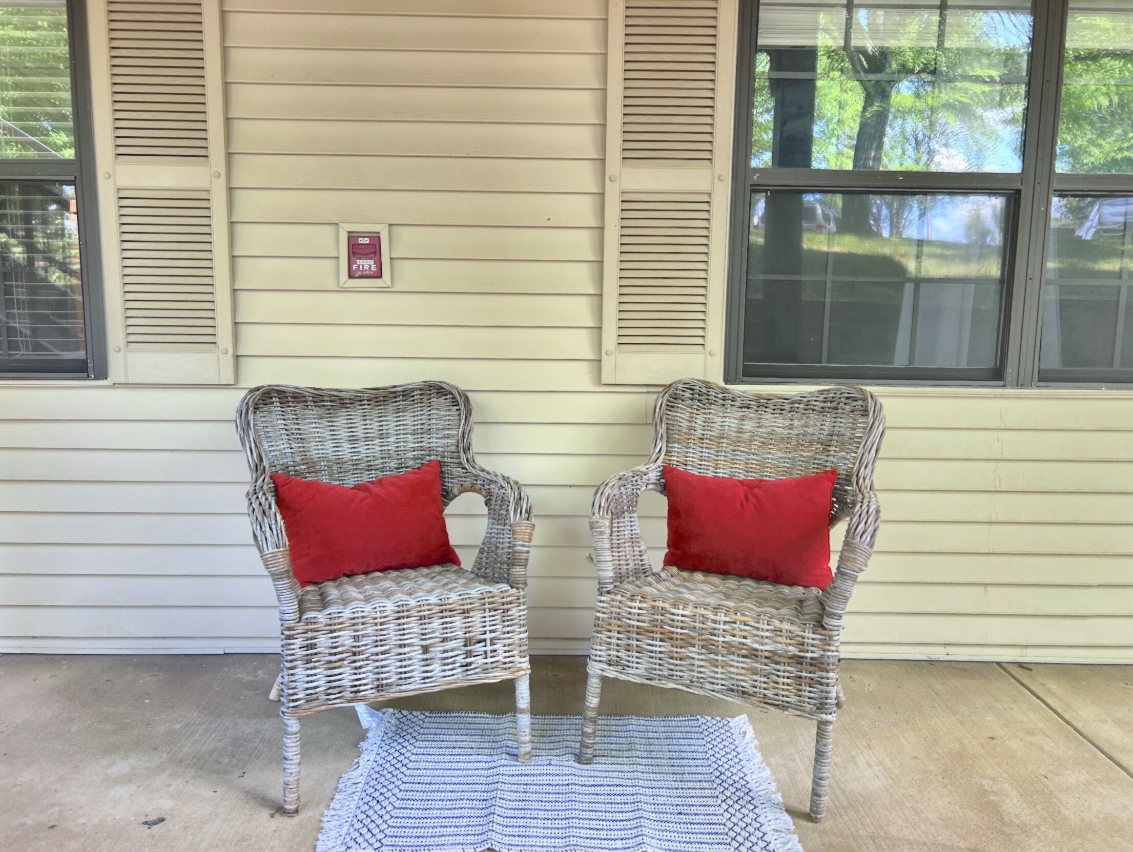 Deux chaises en osier avec des coussins rouges, placées sur un porche doté d’un tapis blanc, devant une maison au revêtement beige et avec un panneau indiquant la présence d’une échelle de secours.