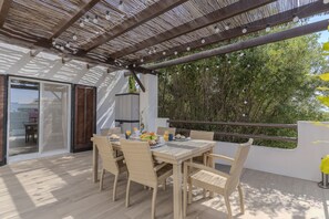 En uteservering med flätade stolar och ett bord dukat för frukost, en pergola ovanför och utsikt över grönskan.