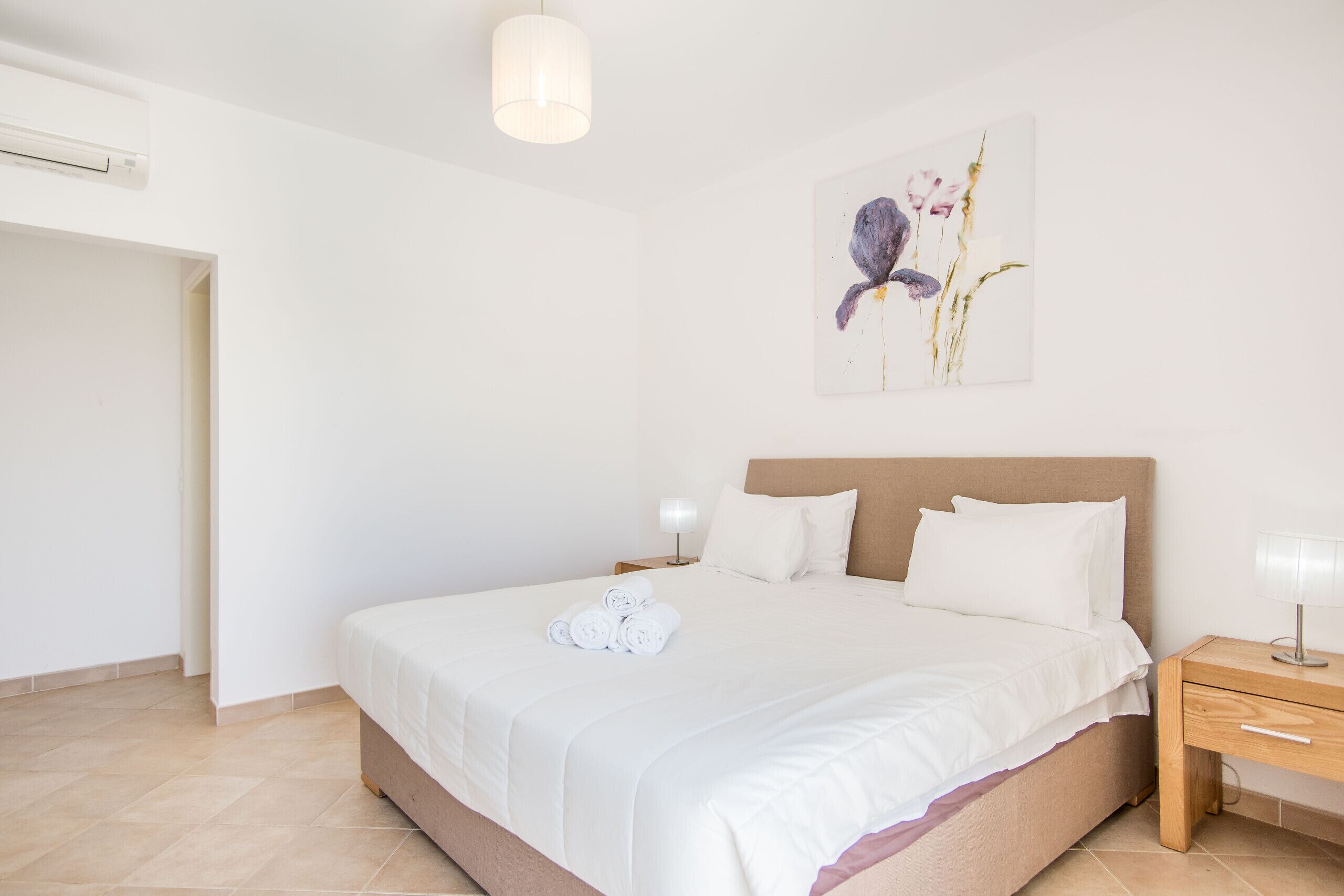Uma cama bem arrumada com lençóis brancos, uma luminária de cabeceira e uma obra de arte na parede.