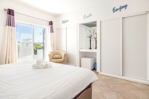 A bedroom with a bed, a chair, a wardrobe, and a view of greenery.