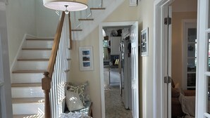 Una escalera de madera con barandal blanco, una lámpara colgante y un tapete que conduce a una zona de dormitorio con cama y sillón.