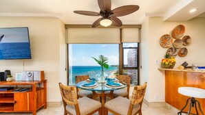 Comedor con vista al océano, ventilador de techo, televisor y encimera de cocina con platos decorativos.