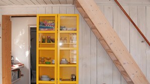 Une pièce avec une étagère jaune contenant divers objets, une petite table surmontée d’un panier en osier et un escalier en bois.
