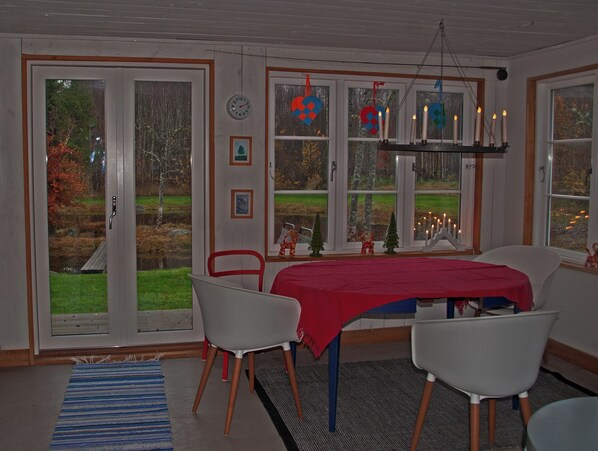 A dining area with a table covered in a red tablecloth, white chairs, and a view of a garden through large windows.
