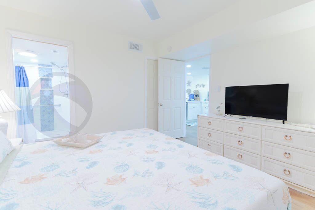 Un dormitorio con cama, cómoda, televisor y baño visible a través de una puerta abierta.