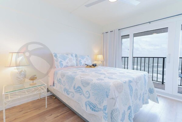 A bedroom with a bed, a nightstand, a lamp, and a view of the sea through large windows.