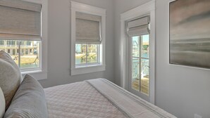 A bedroom with a bed, two windows with blinds, a view of a pool and buildings, and a painting on the wall. - Glass Half Full 205 Fiv-g (Port Aransas)