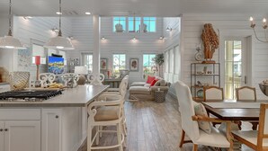 Una cocina amplia con armarios blancos, una isla central e iluminación colgante. Hay un comedor con una mesa y sillas de madera, y un rincón de estar acogedor con un sofá y cojines.