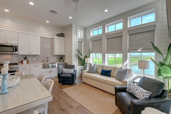 A modern kitchen with white cabinetry, a dining area with a table and chairs, a living room with a sofa and a coffee table, and large windows with blinds.