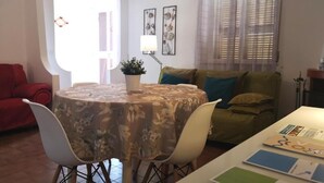 A dining area with a round table covered in a floral tablecloth, surrounded by white chairs. There is a potted plant on the table, and a sofa with cushions is visible in the background. - Appartamento a 100 Metri dal Mare - Tutti i Comfort- Free Wi-fi (Ostuni)