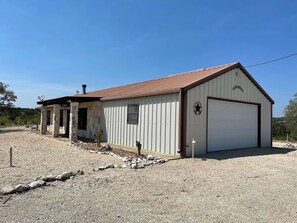 A single-story building with a garage door, a small porch with columns, and a gravel driveway. - Ranchito | Hilltop Cabin w/ Hot Tub & Scenic Views (Hondo)