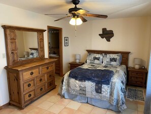 Une chambre avec une commode en bois, un lit avec une literie à motifs, un ventilateur de plafond et un élément décoratif fixé au mur.