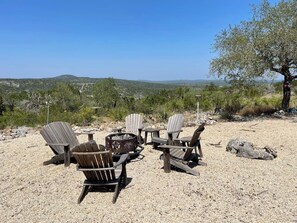 A seating arrangement with Adirondack chairs around a fire pit, set on a gravel surface with a view of distant hills. - Ranchito | Hilltop Cabin w/ Hot Tub & Scenic Views (Hondo)