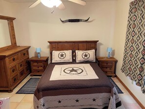 A bedroom with a bed, wooden dresser, nightstands, patterned curtains, and a ceiling fan. - Ranchito | Hilltop Cabin w/ Hot Tub & Scenic Views (Hondo)