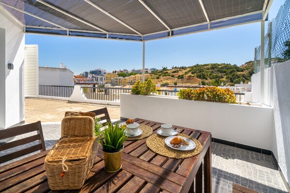 A rooftop terrace with a table set for two, a wicker chair, and a potted plant.