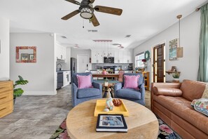 A modern living room with a ceiling fan, two grey sofas with pink cushions, a brown leather sofa, a round coffee table with a tray and a book, and a kitchen in the background. - Family & Dog Friendly Home | Near Beach | Rockets Launch Views | Pool + Kayaks* (Cocoa Beach)