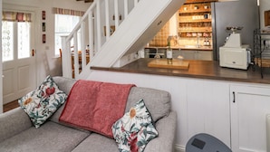 Ein gemütliches Wohnzimmer mit einem grauen Sofa, einer roten Decke und floralen Kissen. Eine Holztreppe führt zu einer Küche mit einer Holz Arbeitsplatte, einem Kühlschrank und einem Spülbecken.