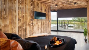 A modern room with a wooden wall, a flat-screen TV, a bed with a black bedspread, and a dining table with a tray of food. - Rusty Boathouse (Llanarth)
