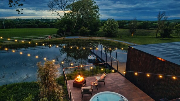 A hot tub on a patio with string lights, surrounded by a wooden deck and a view of a pond. - Rusty Boathouse (Llanarth)