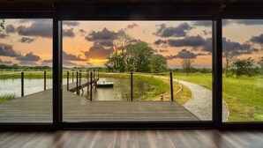 A modern interior with large windows offering a view of a sunset over a water body and a grassy landscape. - Rusty Boathouse (Llanarth)