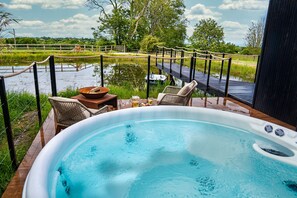 Un jacuzzi con terraza de madera y sillas, con vista a un río y un puente de madera.
