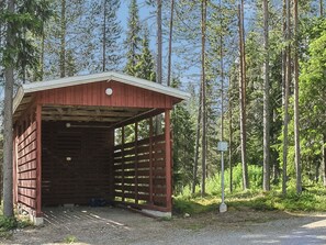 Um pequeno abrigo de madeira com telhado inclinado, entrada de cascalho e um poste de metal em uma área arborizada.