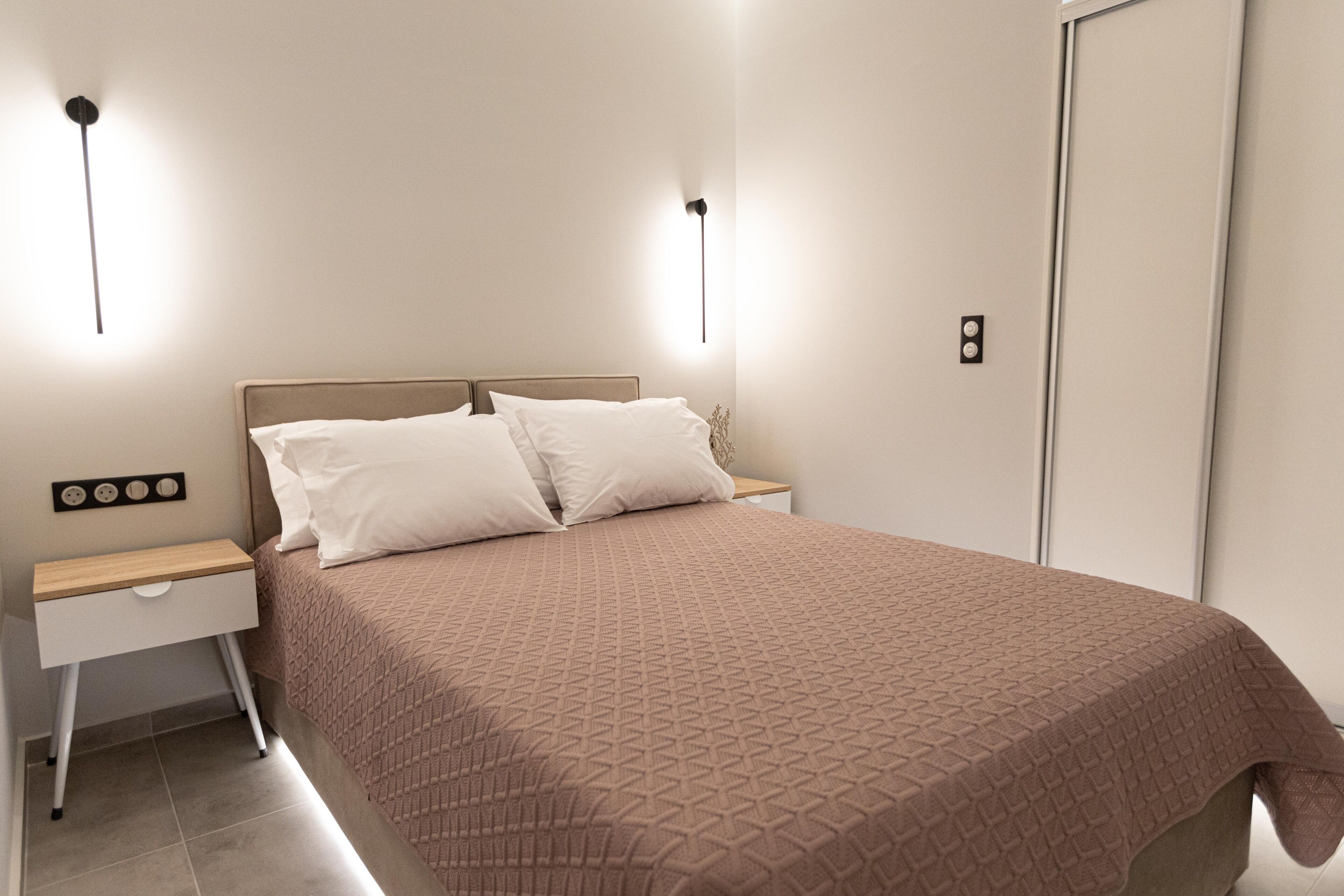 Una cama bien hecha con una colcha estampada, dos mesitas de noche con lámparas y un enchufe en la pared.