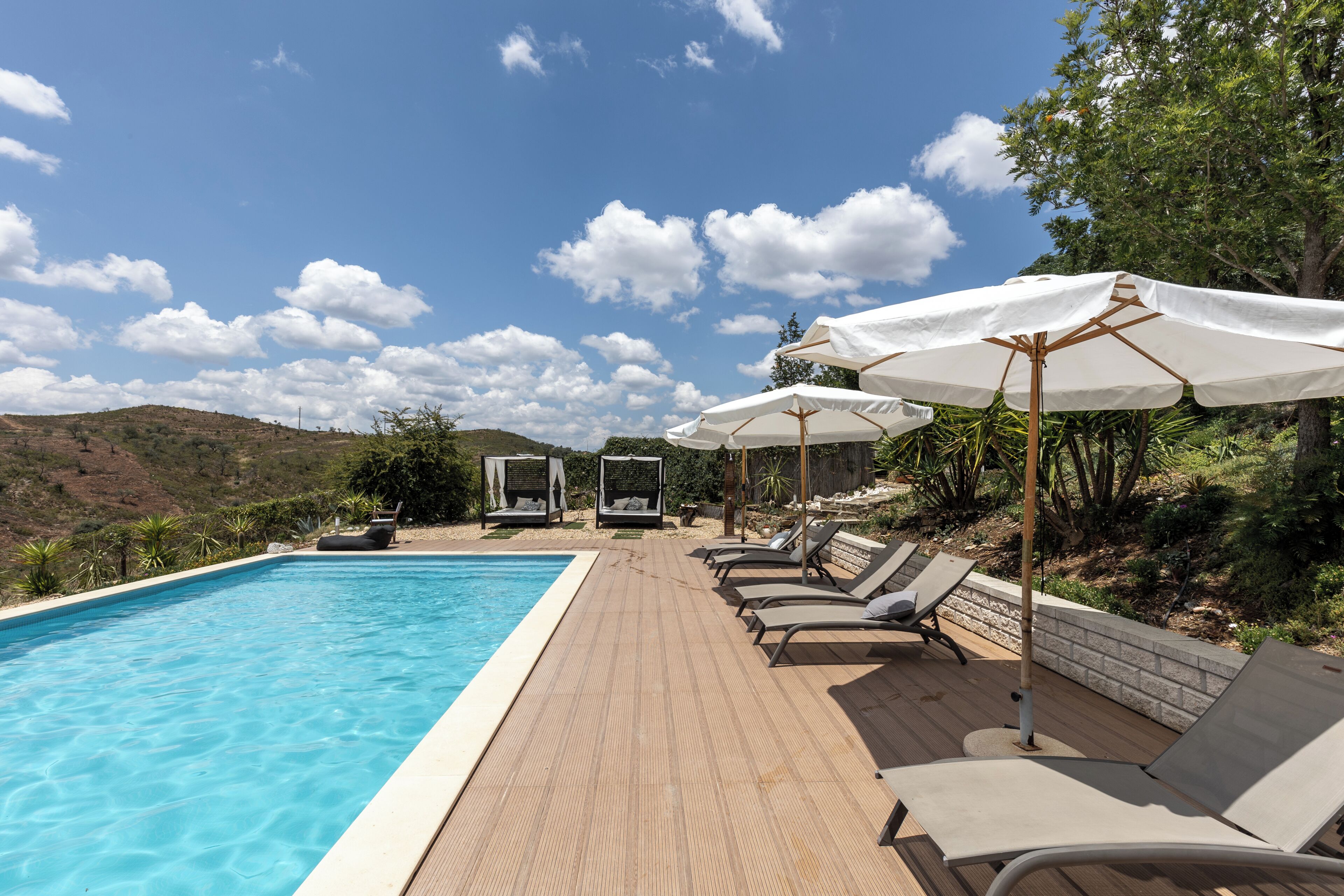 A poolside area with lounge chairs and umbrellas, a clear sky, and a hilly landscape in the background.