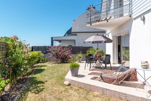 Une maison moderne dotée d’un patio comprenant une table, des chaises, une chaise longue et un pot de fleurs.