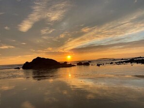 Un coucher de soleil sur une plage, avec une grande formation rocheuse au premier plan.