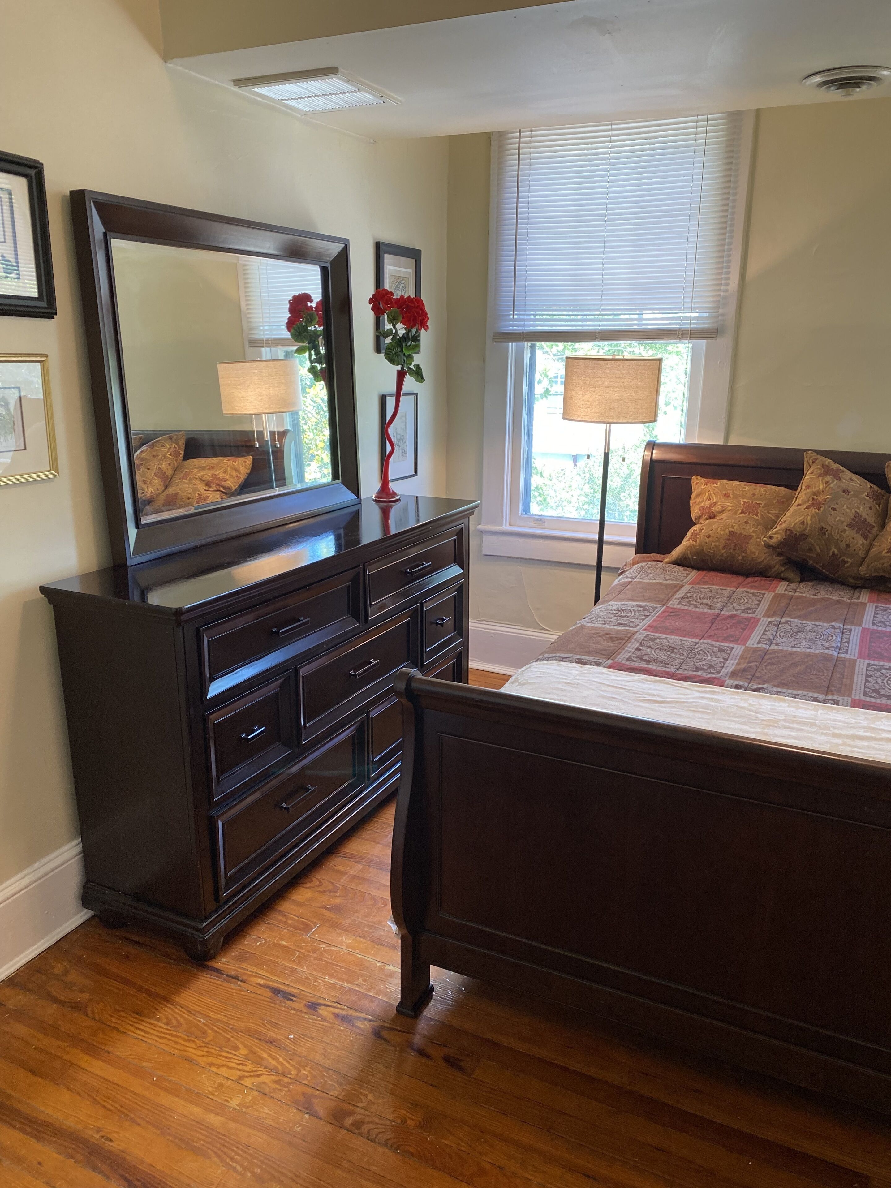 Een slaapkamer met een houten dressoir, een grote spiegel, een bed met beddengoed met een patroon en een raam met zonwering.