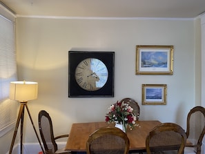 Un comedor con una mesa de madera, sillas de mimbre, un reloj enmarcado y dos cuadros en la pared.