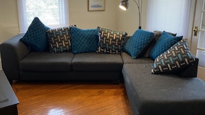 A living room with a dark-colored sectional sofa, patterned cushions, a floor lamp, and two framed pictures on the wall.