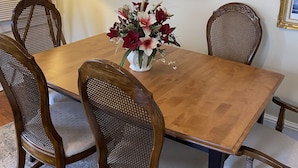 A dining area with a wooden table, four wicker chairs, a wall-mounted clock, and a vase with flowers.
