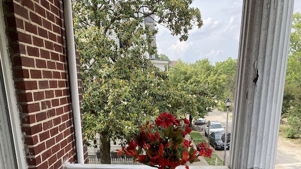 A glass-top table with two cushioned chairs on a wooden deck, a vase with red flowers, and a brick wall.