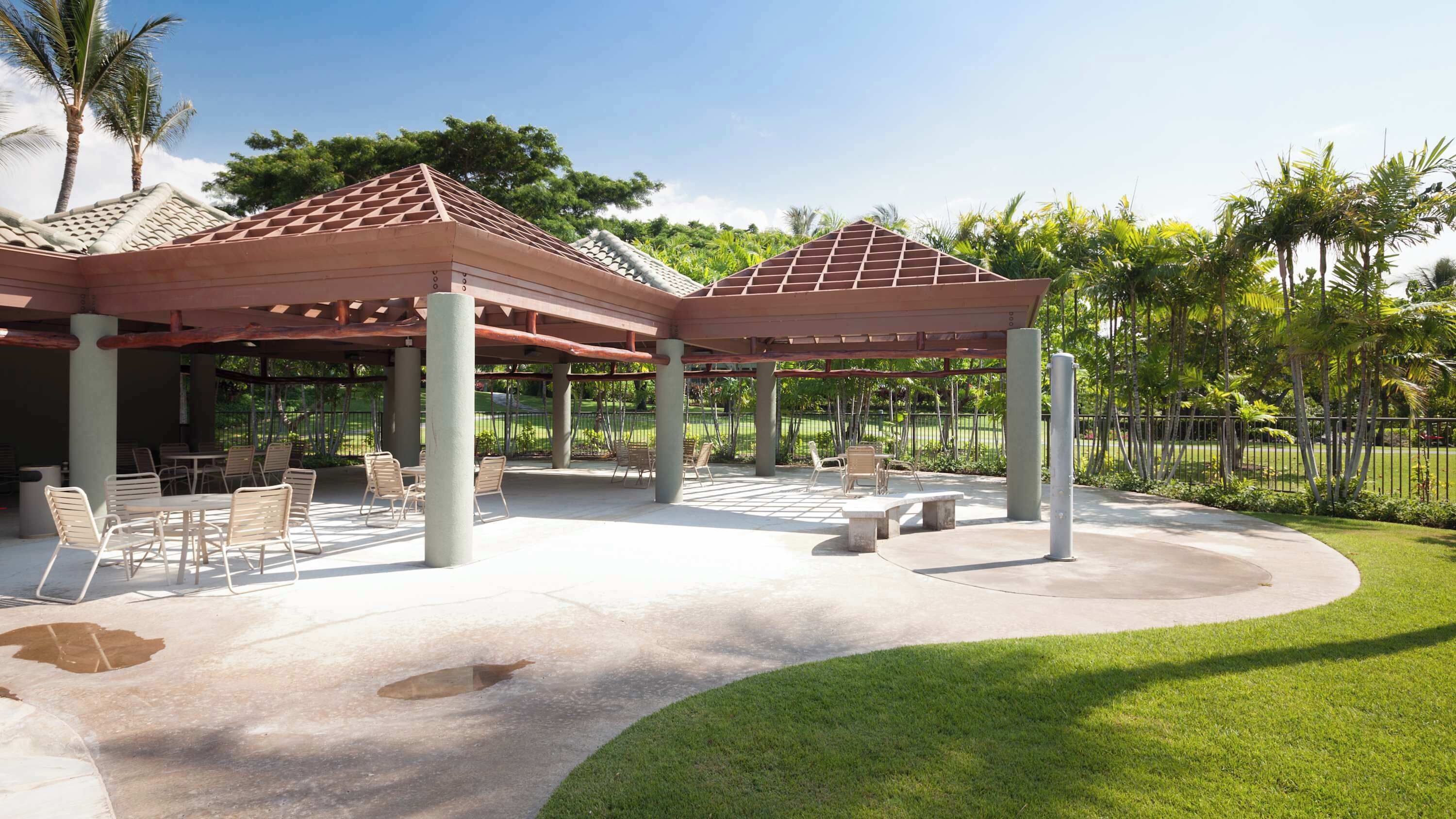 A covered outdoor patio with tables and chairs, surrounded by a well-maintained lawn and palm trees.